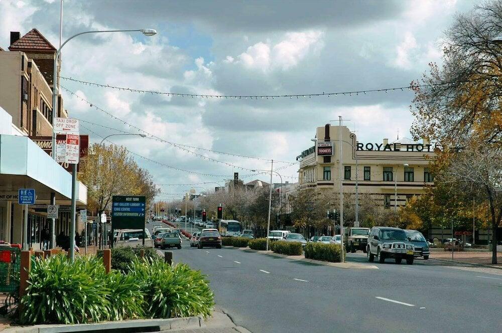 View down a town's main street featuring the prominent Royal Hotel on the right, with cars, shops, trees, and overhead string lights under a partly cloudy sky.