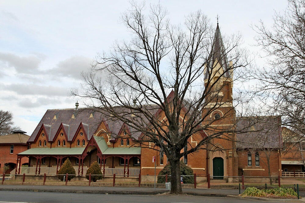 Large historic red brick building with a steeple and gabled roof, partially obscured by a large bare tree in winter.